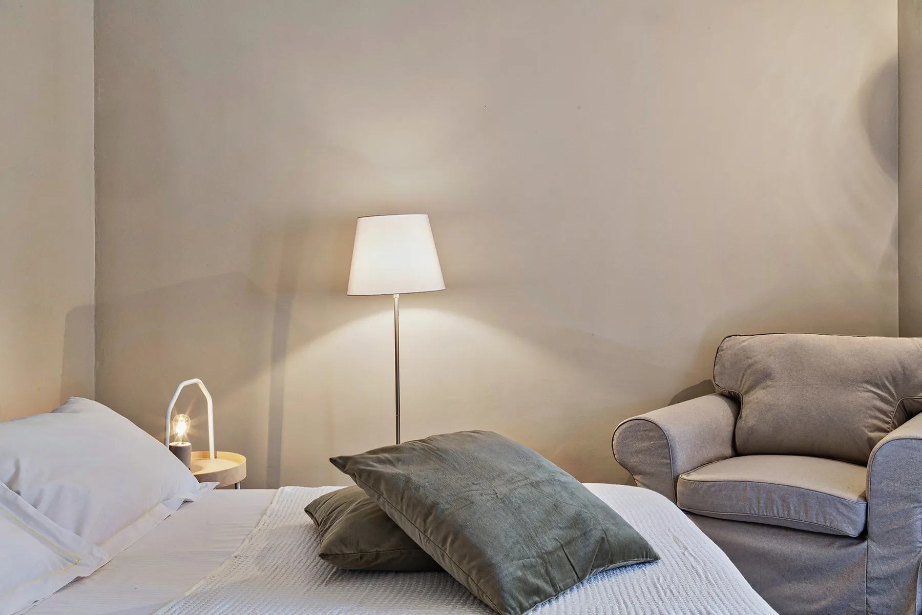 Dormitorio con cama, almohadas y una lámpara de pie junto a asientos tapizados.