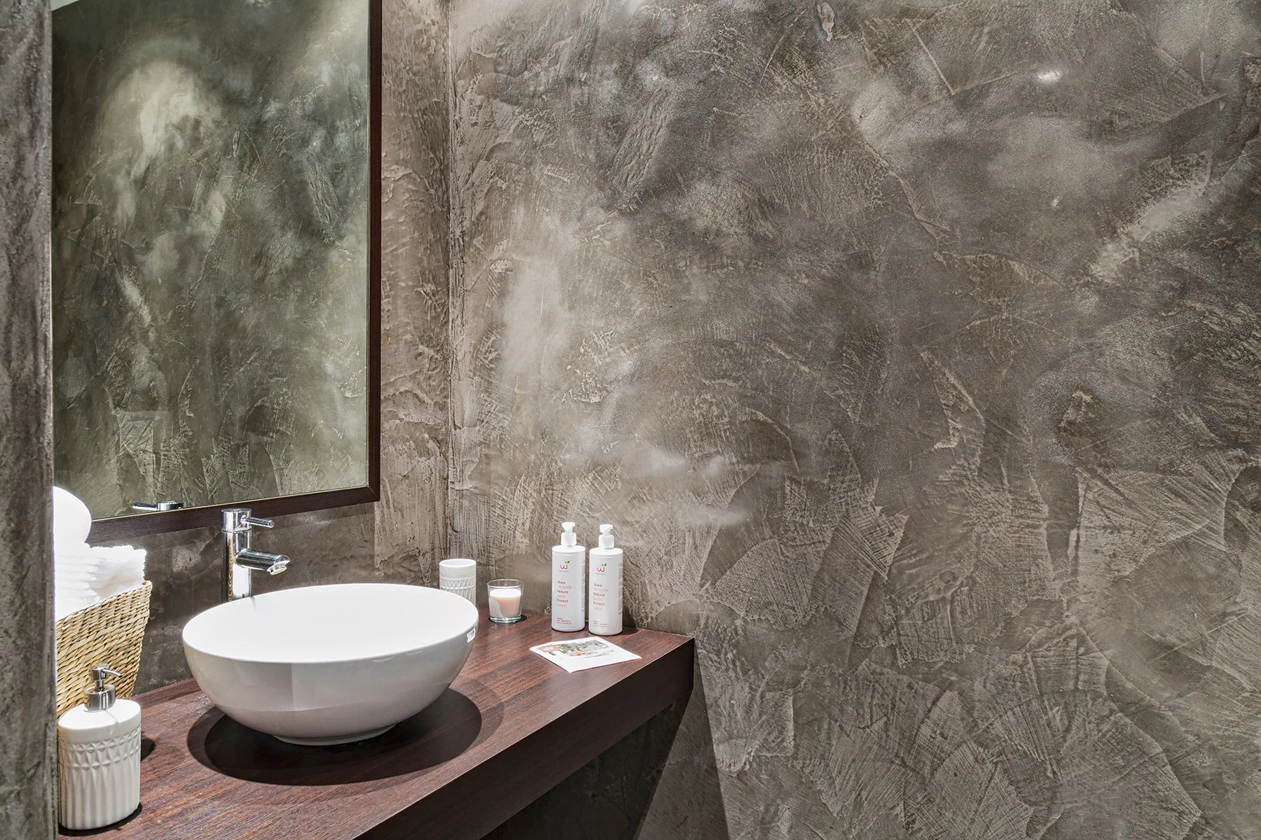 Un tocador de baño con lavabo blanco sobre encimera y artículos de aseo junto a una pared texturizada.