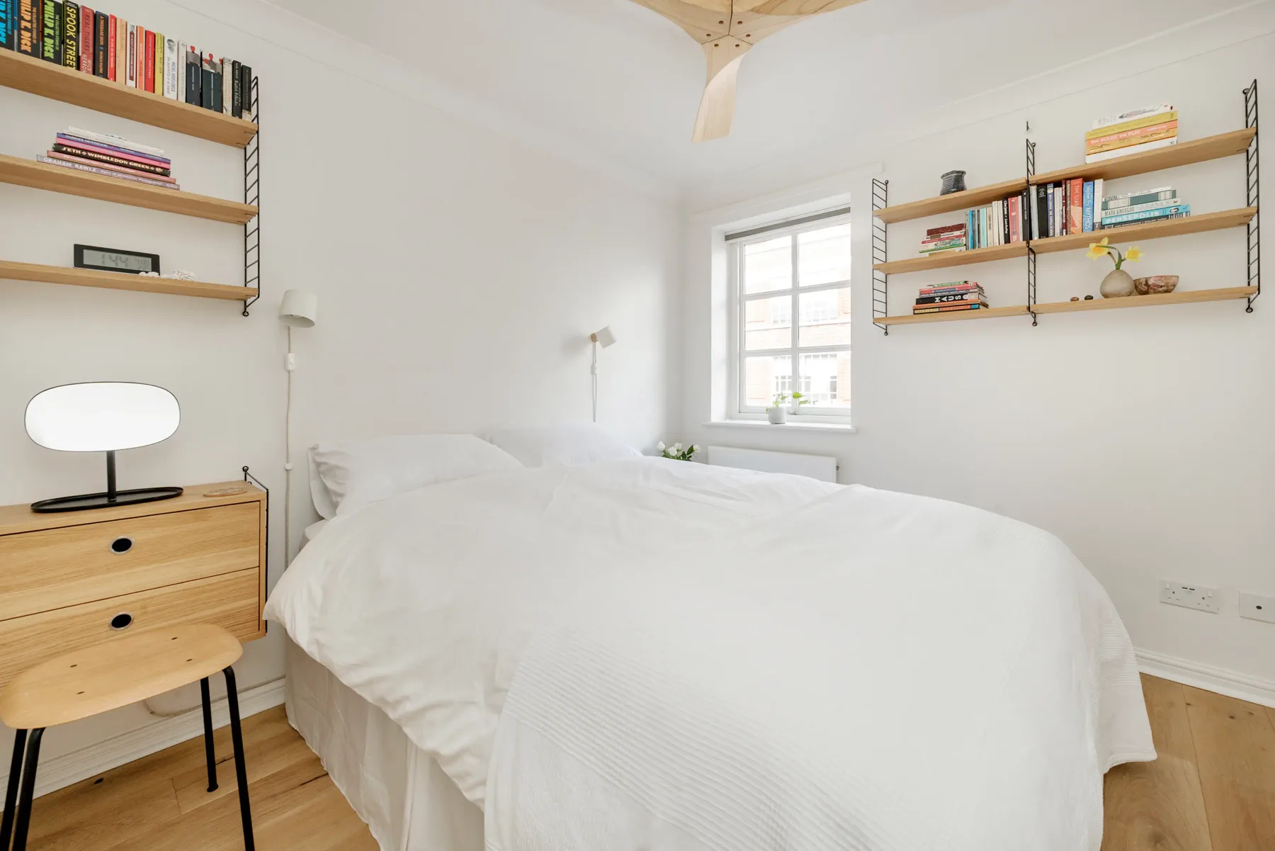 A bright bedroom with a white bed, wooden shelves, and a window.