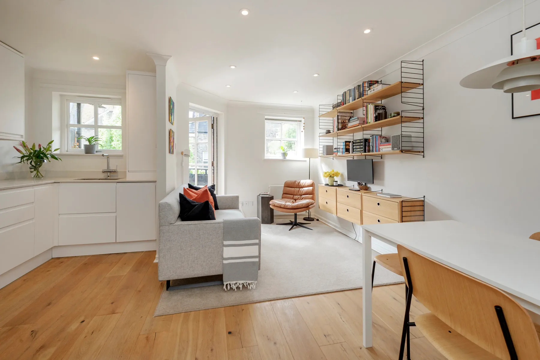 A bright open-plan living area with a sofa, desk, and wall-mounted shelves.