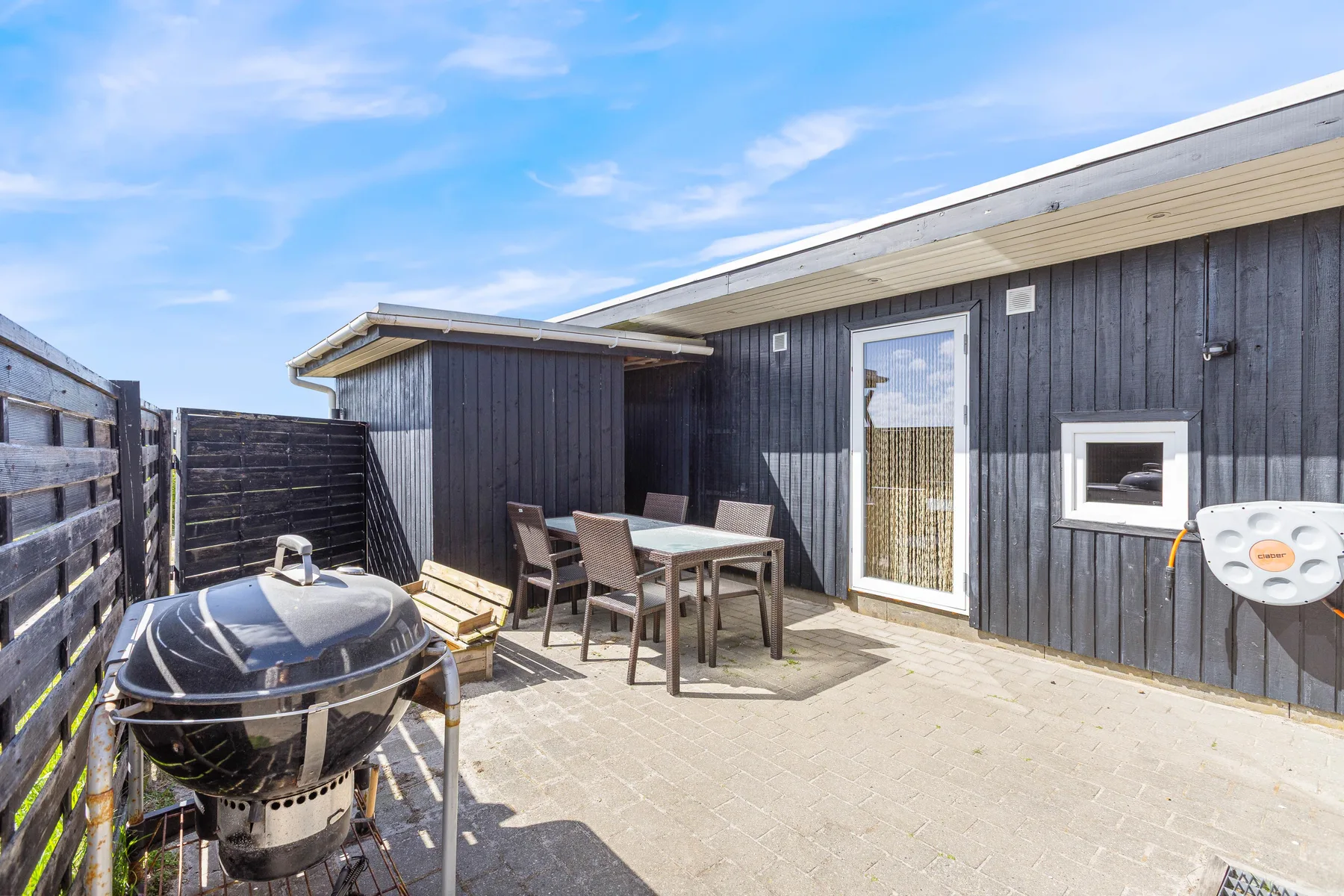 An outdoor patio with a dining table and barbecue beside a dark-sided house.