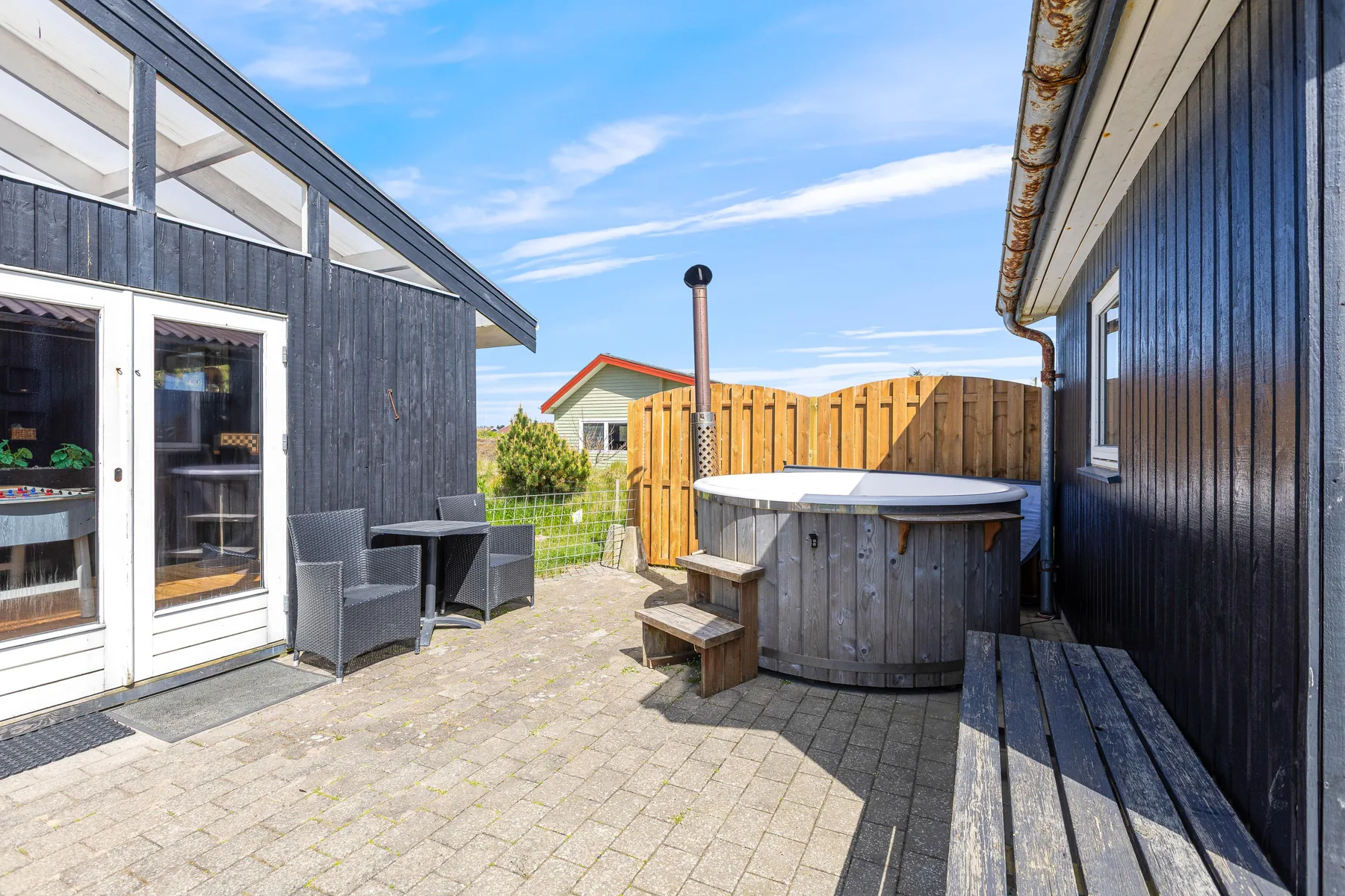 A paved outdoor patio with a hot tub, seating, and a wooden fence.