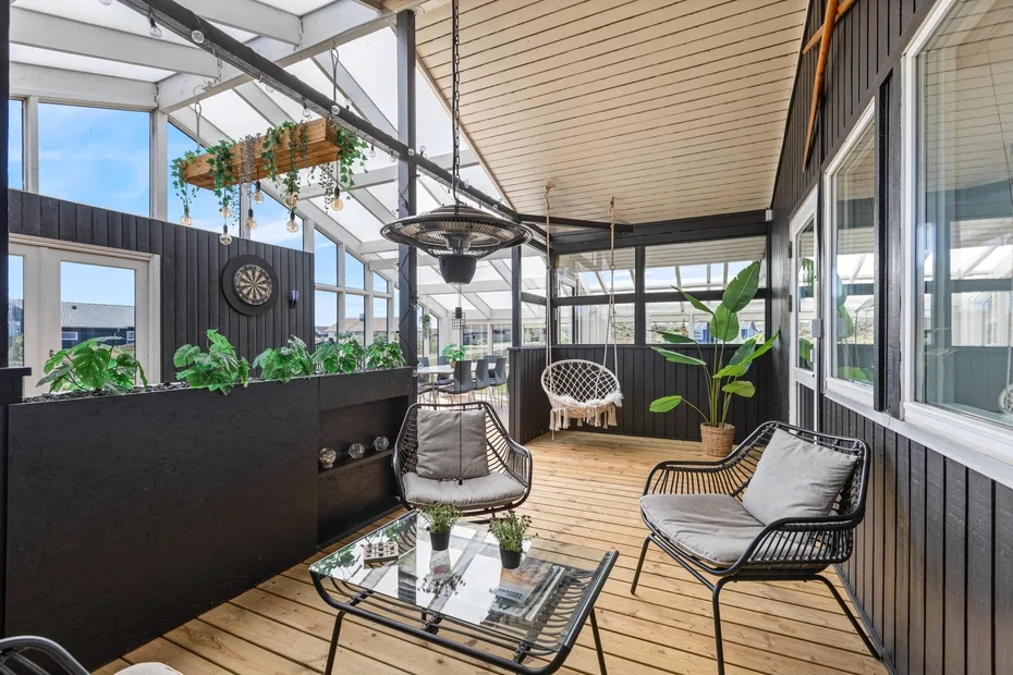 A glass-enclosed sunroom has lounge seating, potted plants, and a wooden deck.