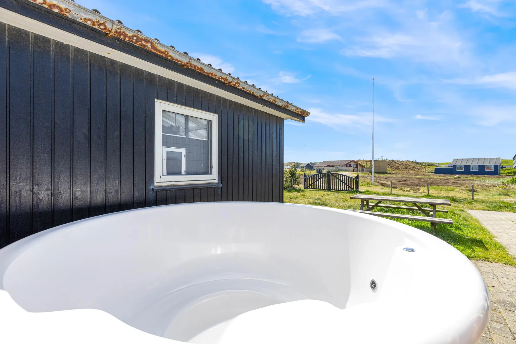 An outdoor hot tub sits beside a dark-sided house, with open dunes beyond.