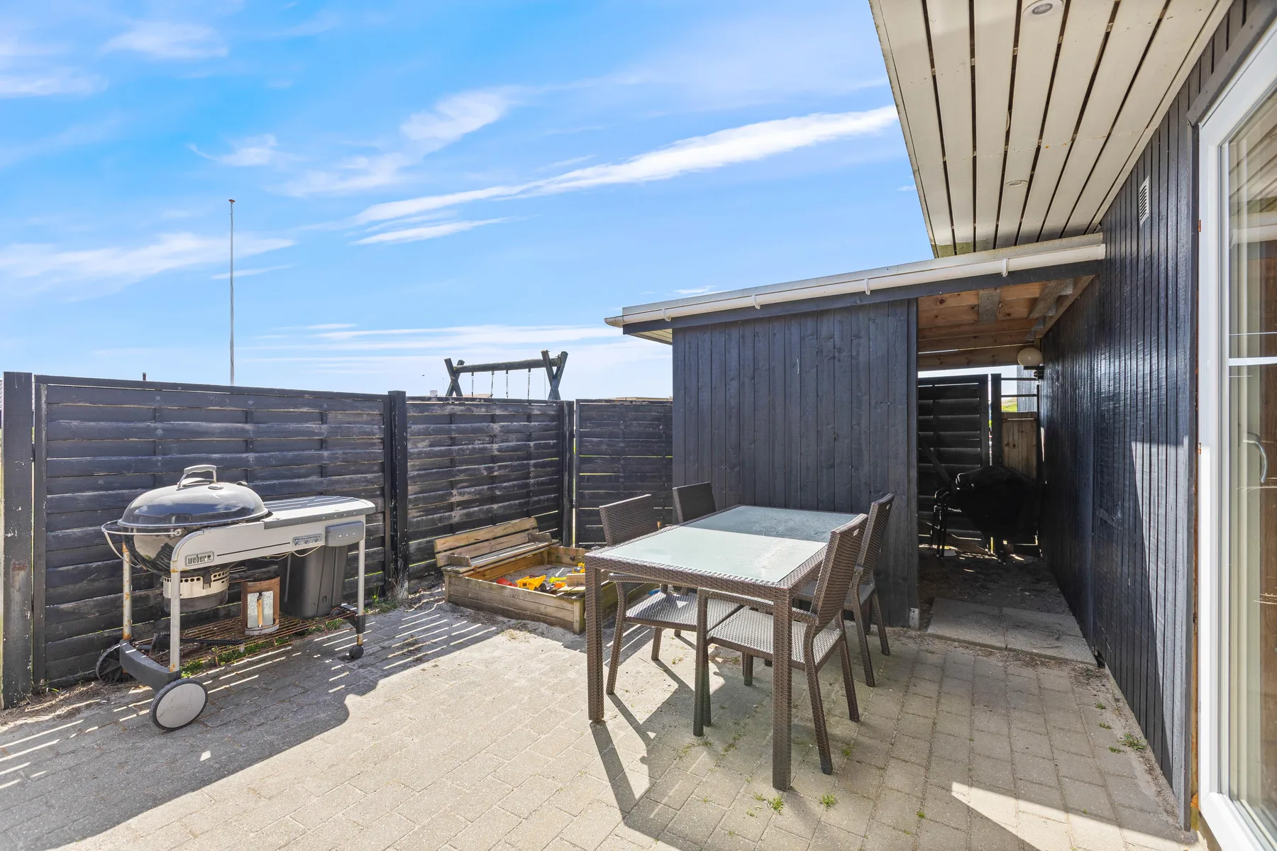 A paved outdoor patio with a dining table, seating, and a grill beside a dark wooden fence.