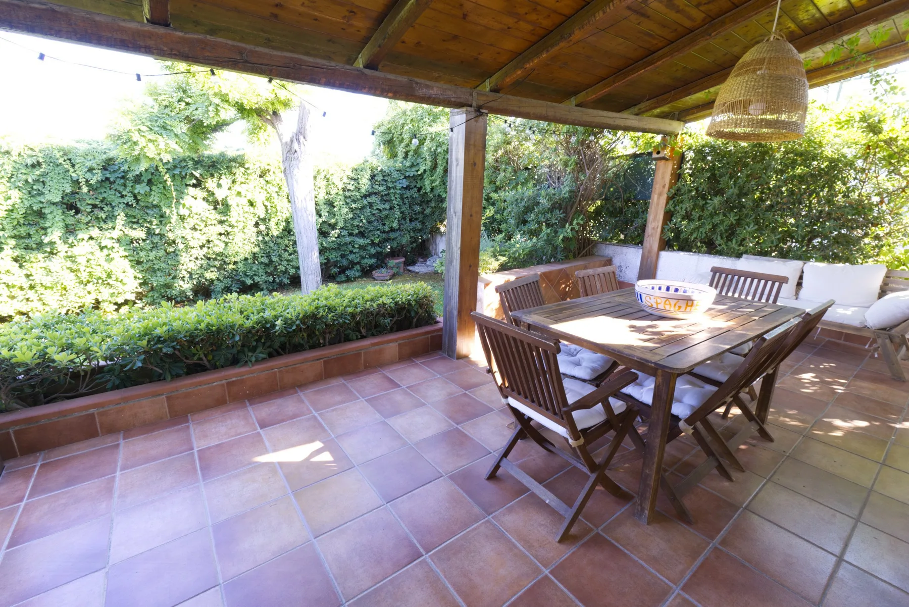 A covered outdoor dining area with a wooden table and surrounding greenery.