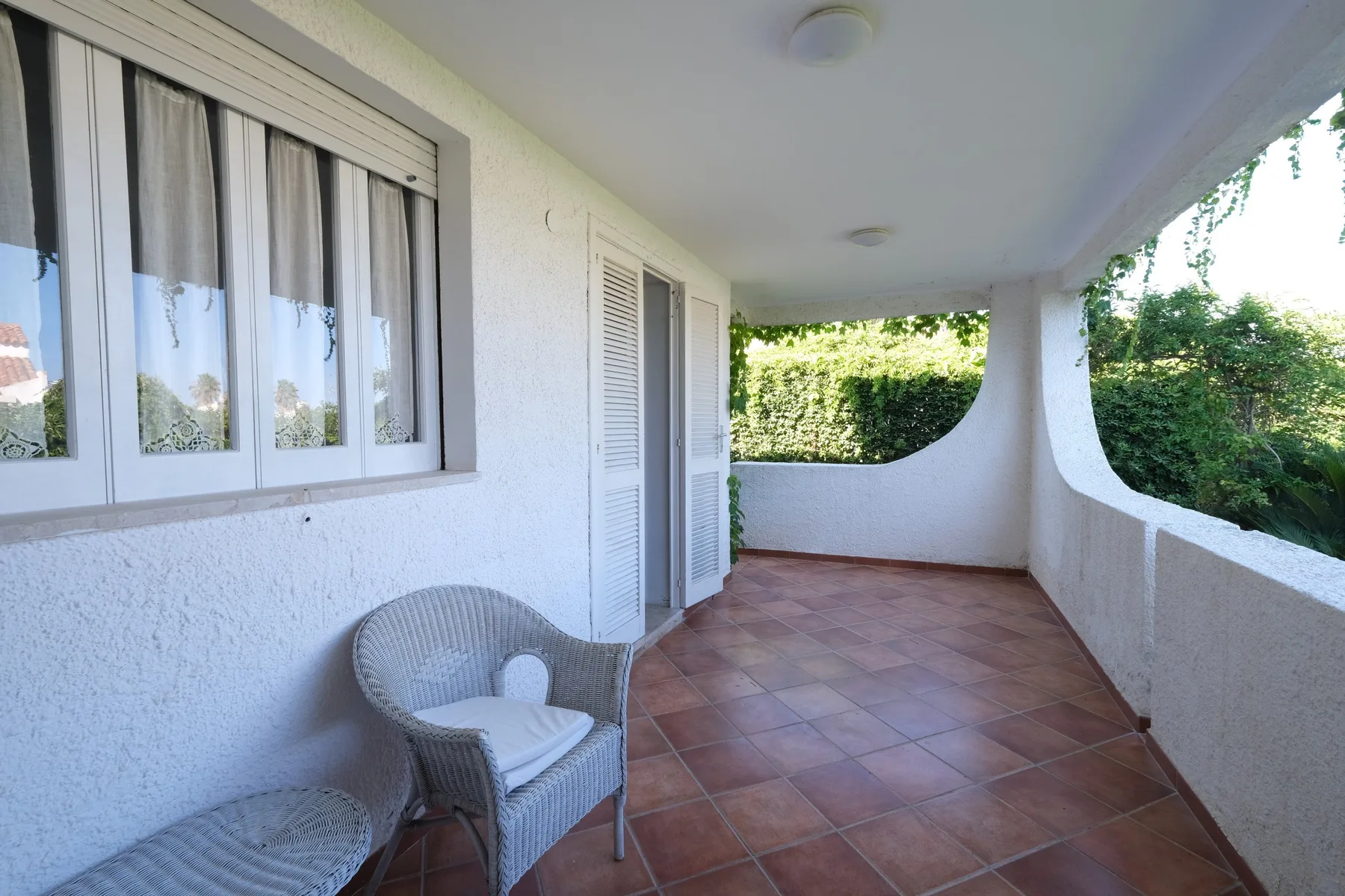 A covered outdoor patio with tiled flooring and seating beside a window.