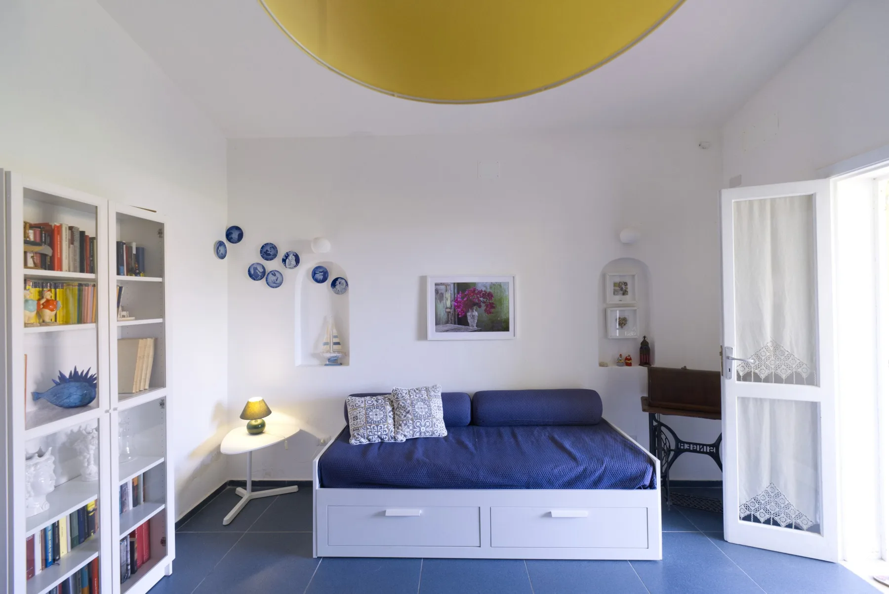 A bright bedroom with a blue bed, white storage shelves, and an open balcony door.