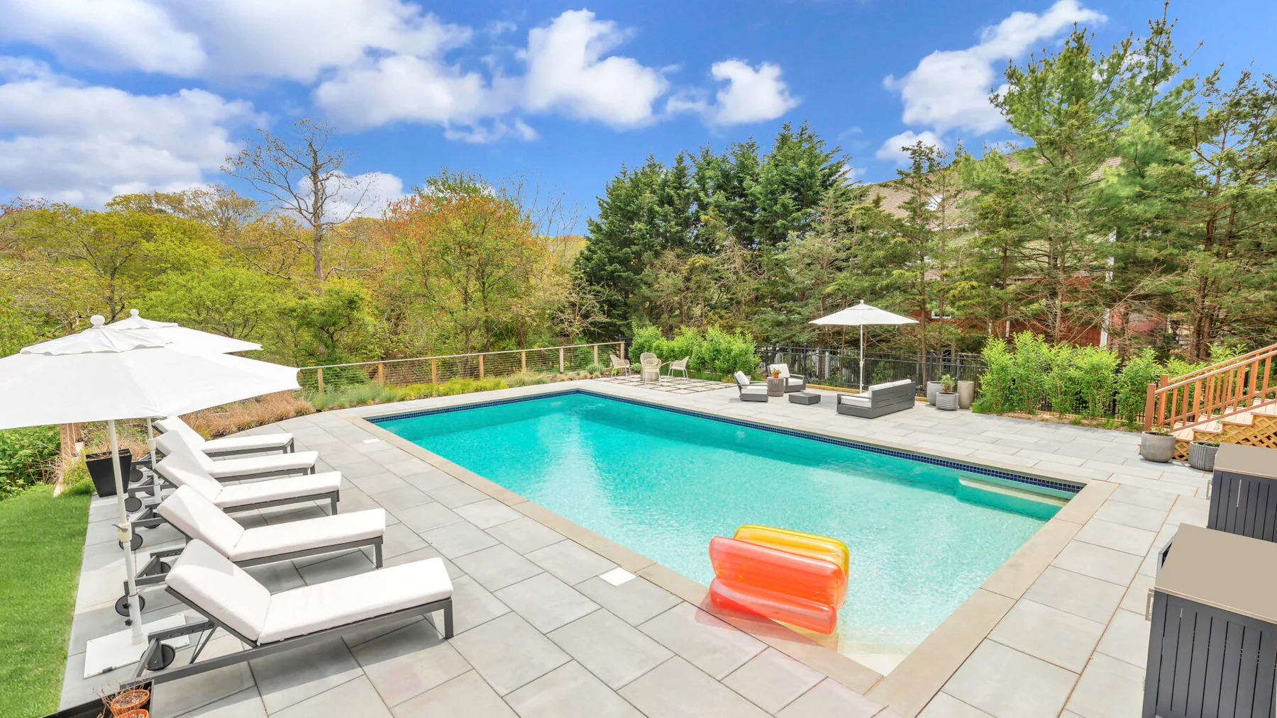 Una piscina está junto a un patio pavimentado con asientos reclinables y sombrillas.