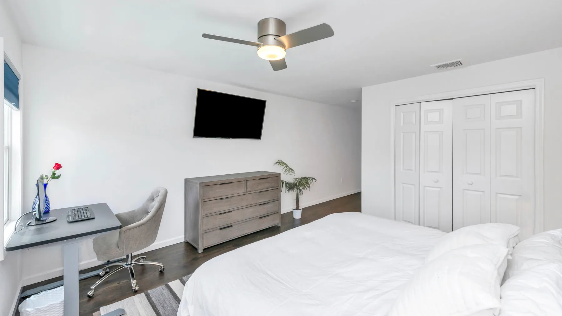 Un dormitorio con una cama blanca, un escritorio y un televisor en la pared.