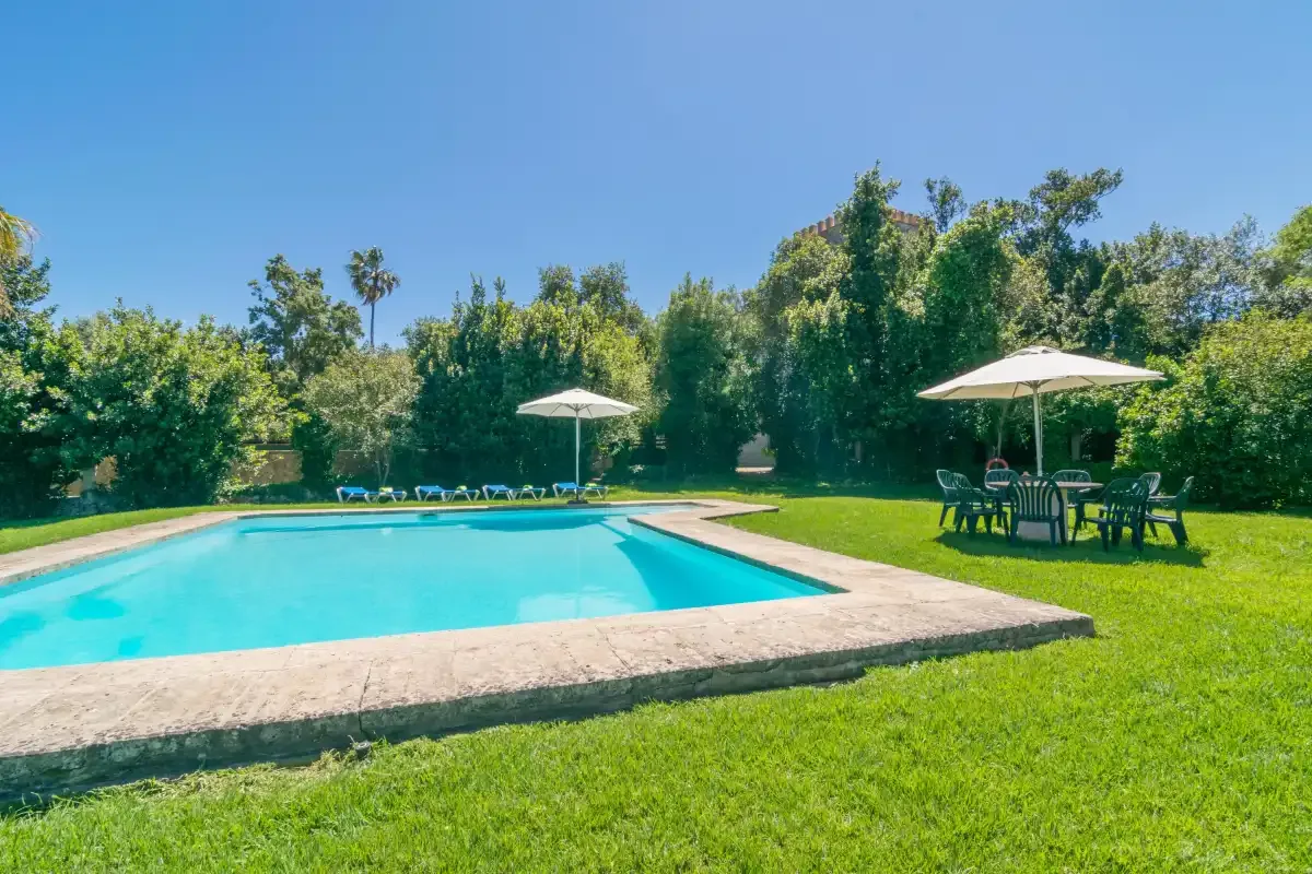 Una piscina al aire libre está rodeada de césped, árboles y asientos a la sombra.