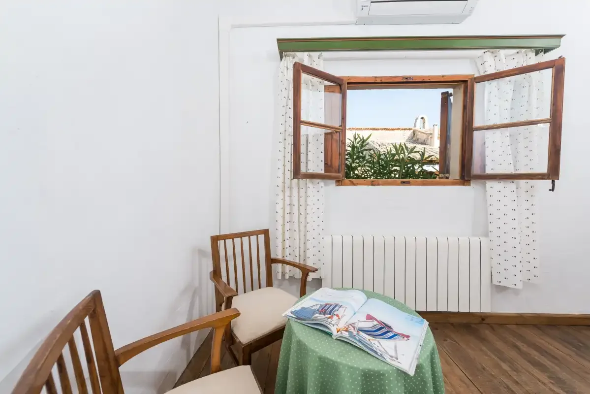 Una habitación con asientos de madera, una mesa con mantel verde y una ventana abierta.