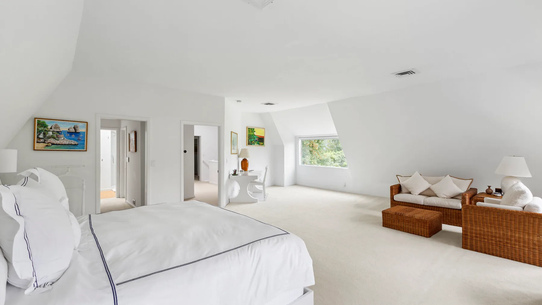 Un dormitorio luminoso con una cama blanca y una pequeña zona de estar junto a la ventana.