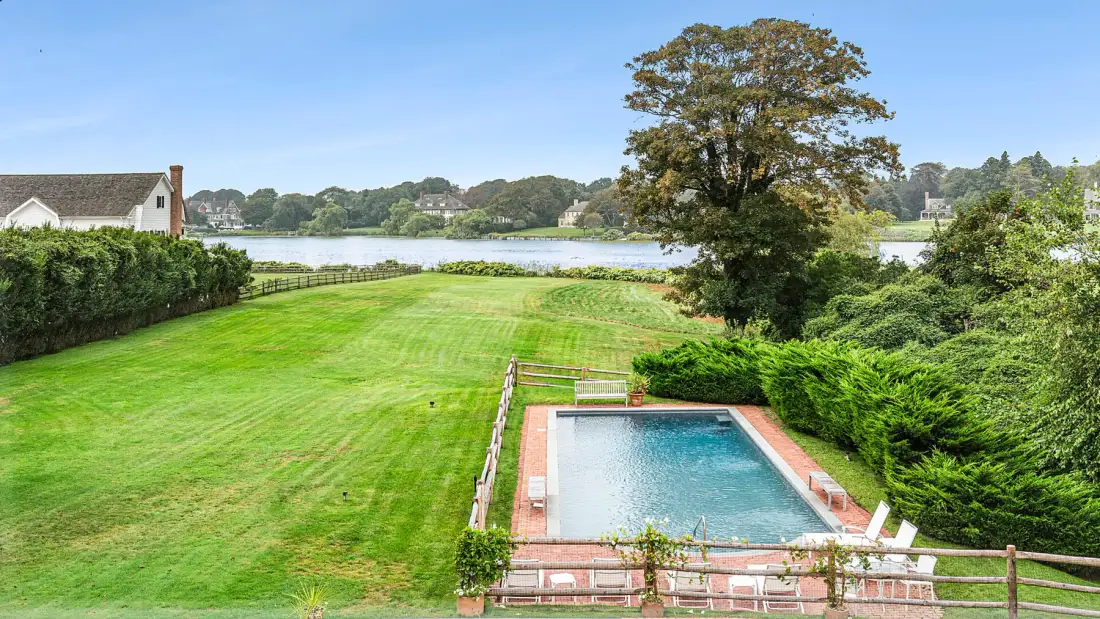 Una piscina y un césped verde miran hacia un lago bajo un árbol grande.