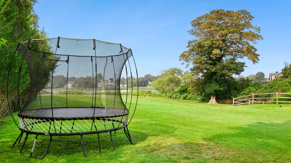 Un trampolín está en un amplio césped junto a un árbol grande.