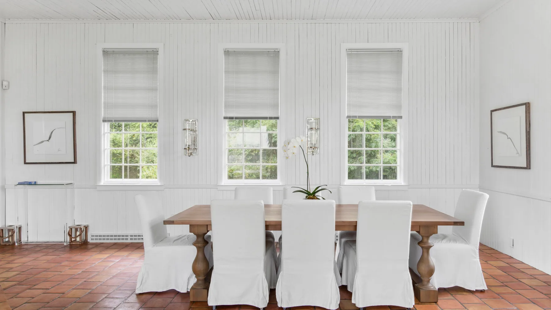 Un comedor luminoso con una mesa larga de madera y asientos cubiertos de blanco.