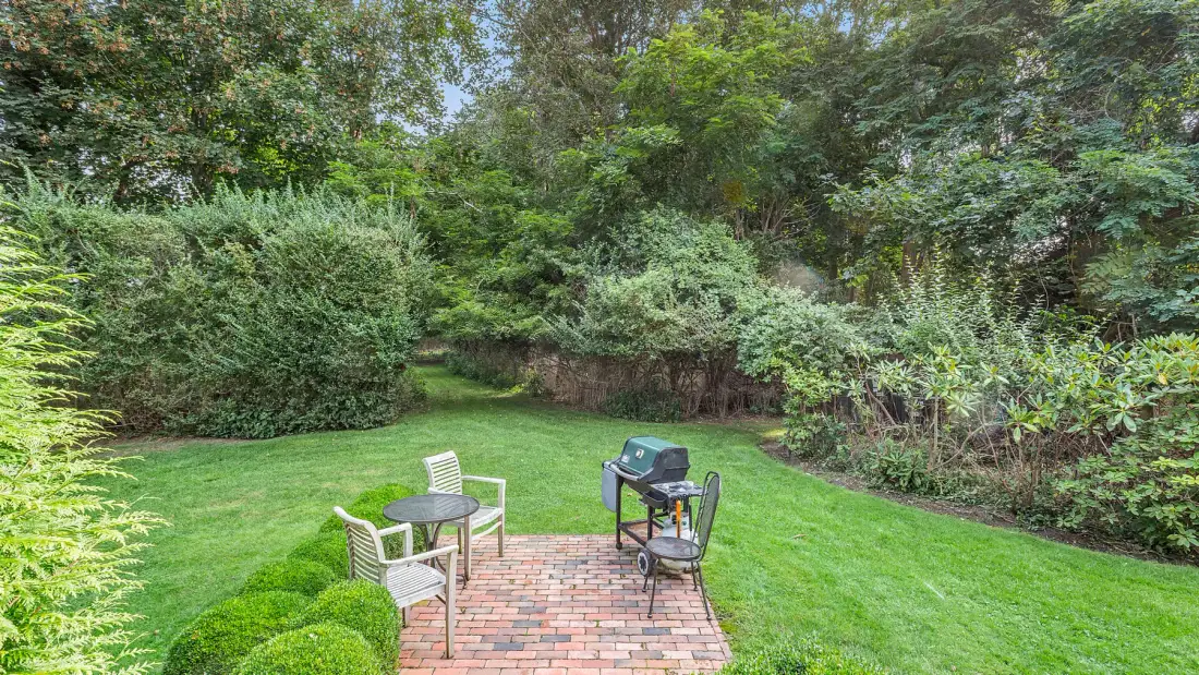 Un patio de jardín con asientos al aire libre está junto a un césped y abundante vegetación.