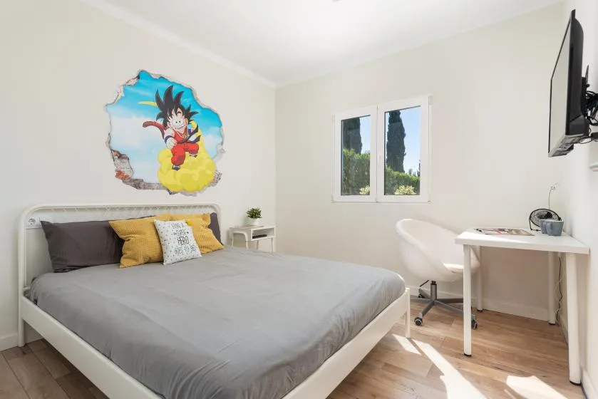 A bedroom with a neatly made bed, wall art, and a desk beside the window.