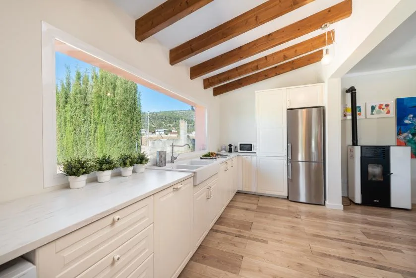 A bright kitchen with white cabinets, a long counter, and exposed wooden ceiling beams.