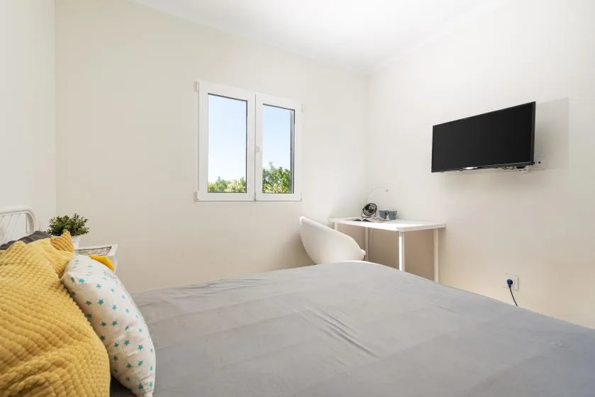 A bright bedroom with a bed, a desk, and a wall-mounted television.