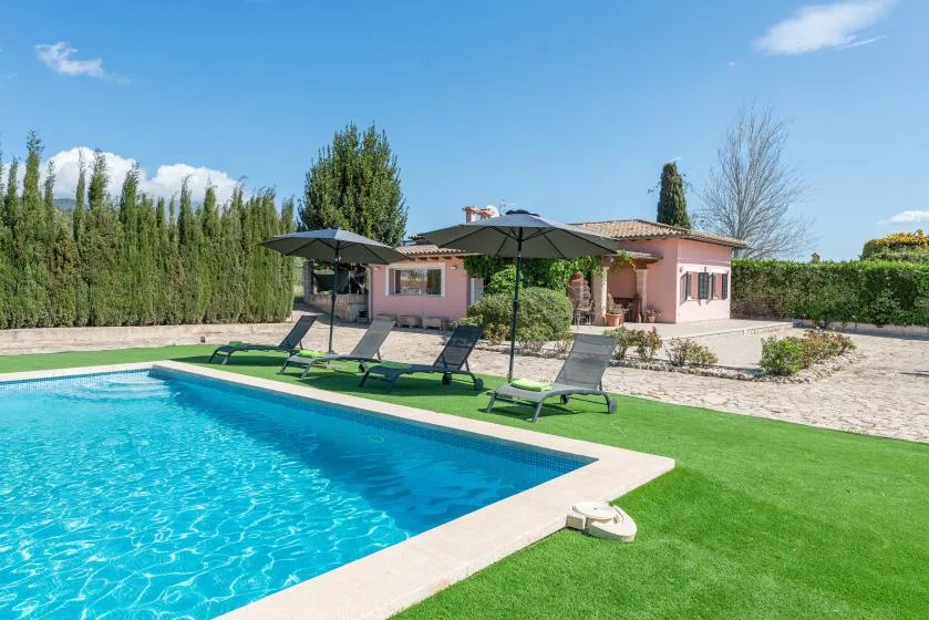 An outdoor swimming pool beside a pink house, with loungers on the lawn.