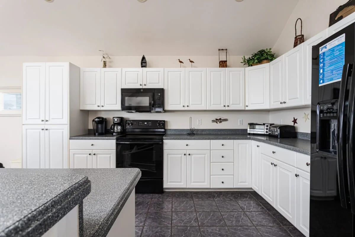 Una cocina luminosa con gabinetes blancos, encimeras oscuras y un refrigerador grande.