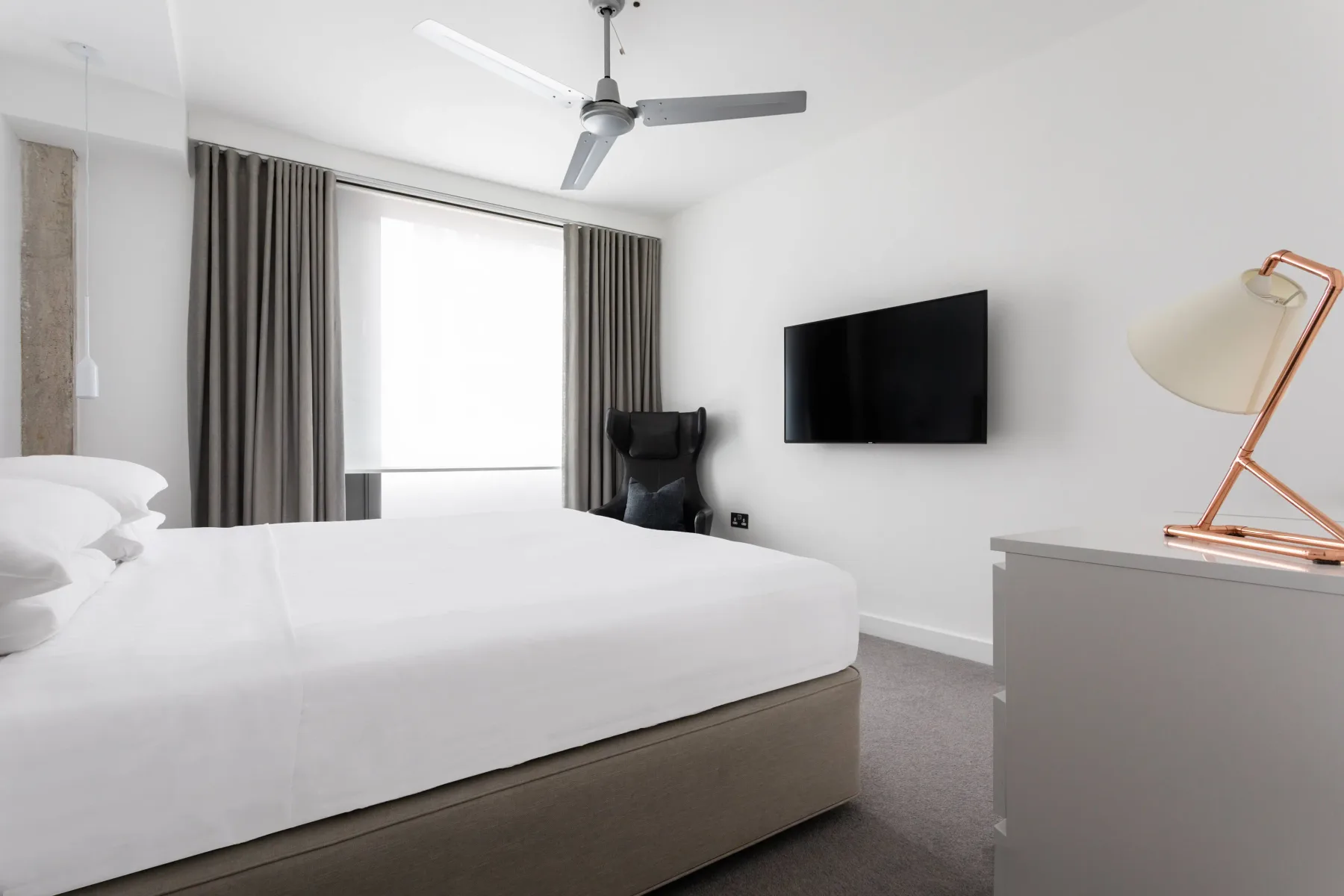 A bright bedroom with a large bed, curtained window, and wall-mounted television.