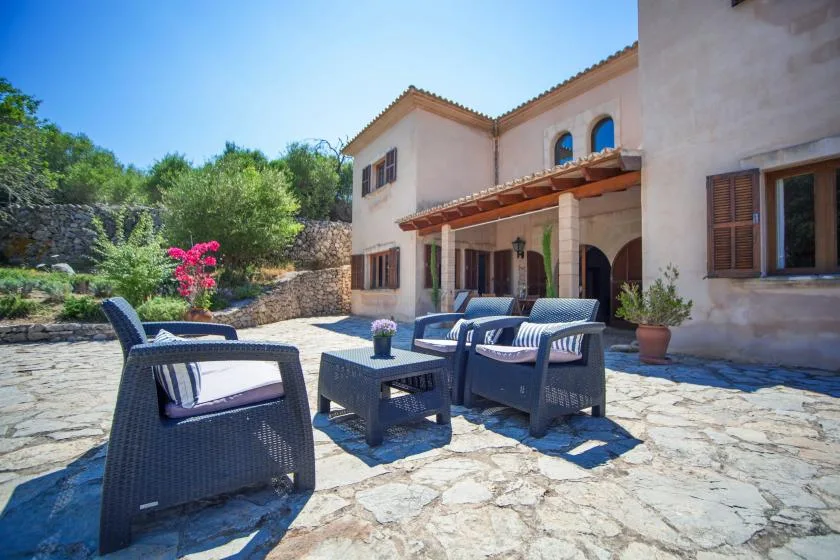 A stone patio with outdoor seating sits beside a stucco house and garden.