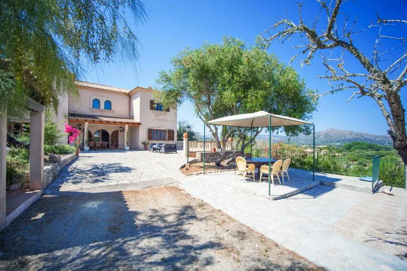 A sunny outdoor patio beside a house, with a dining table and chairs under a canopy.