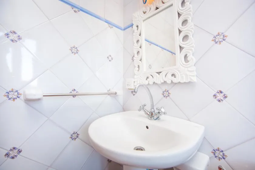 A bathroom sink sits beneath an ornate mirror against tiled walls.