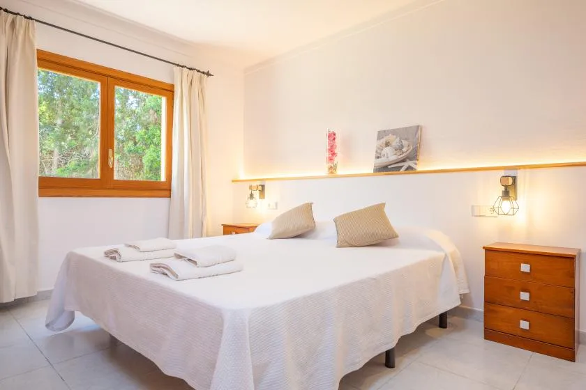 A bedroom with a neatly made bed, wooden-framed window, and bedside chest.