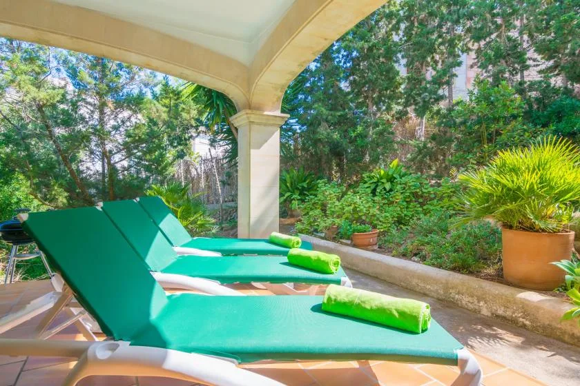A covered outdoor lounging area with green reclining chairs beside leafy garden plants.
