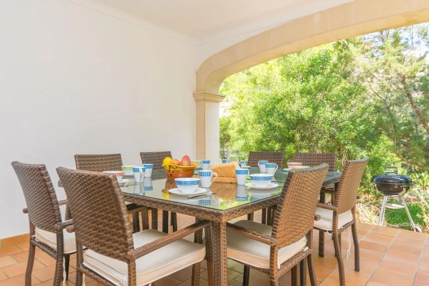 A covered outdoor dining area has a table and seating beside leafy trees.