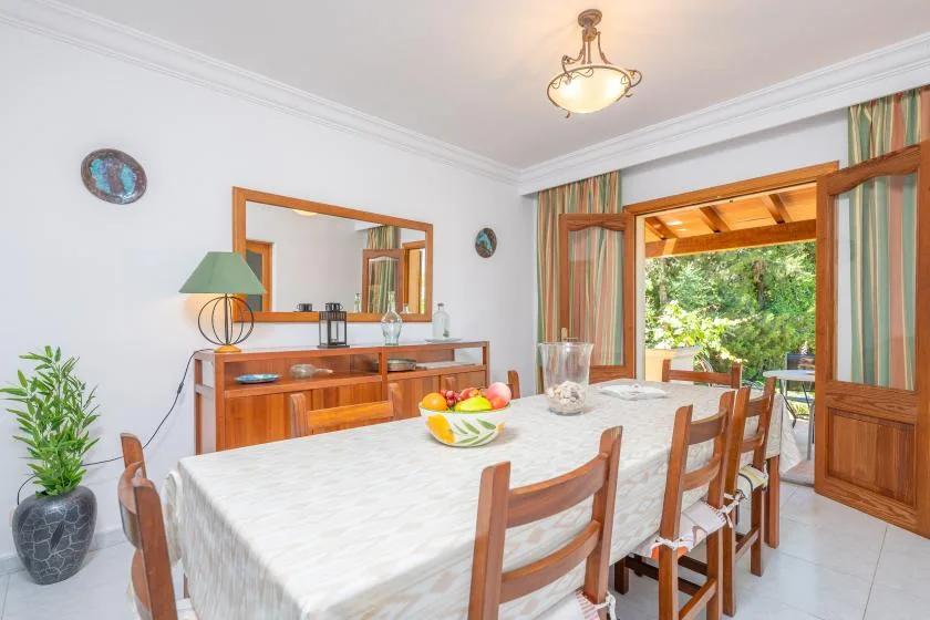 A dining room with a long table set beside open wooden doors to a leafy patio.