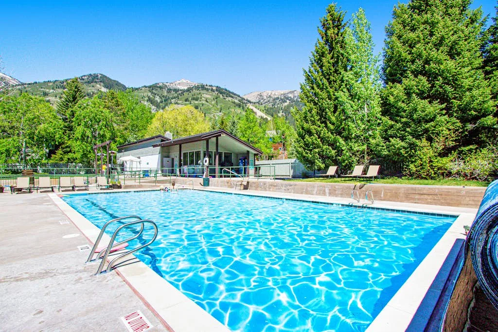 An outdoor swimming pool sits beside a low building, with trees and hills beyond.