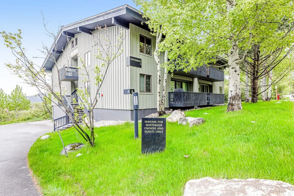A modern building with dark balconies stands beside a green lawn and trees.