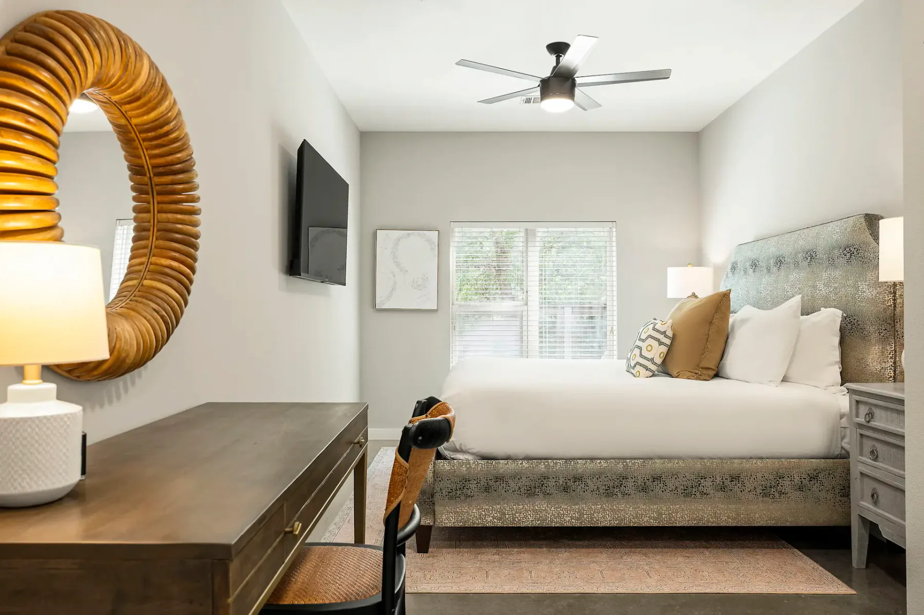 Dormitorio moderno con una cama grande, cojines decorativos, un escritorio de madera, una silla, un espejo redondo, un televisor montado en la pared y un ventilador de techo. Luz natural procedente de la ventana.