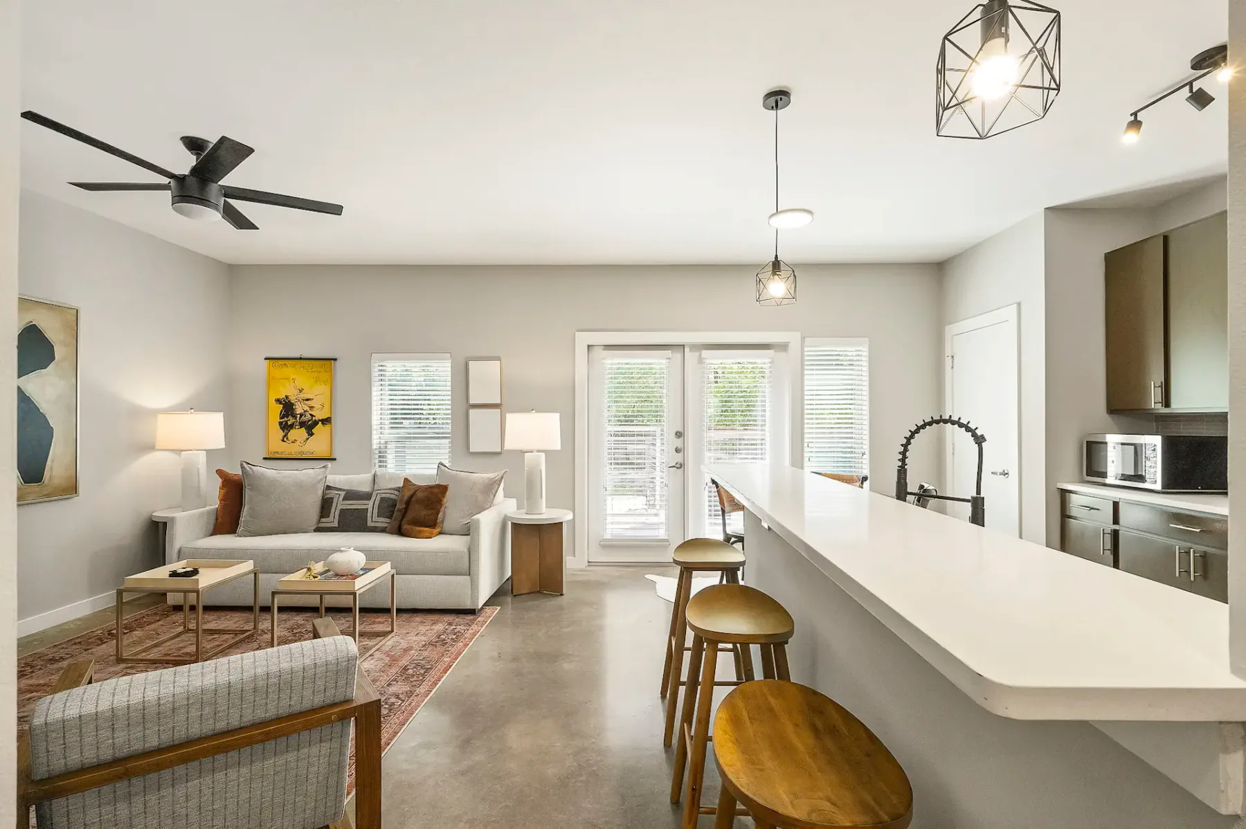 Zona de estar moderna de planta abierta con un sofá acogedor, una isla de cocina con taburetes, lámparas colgantes, ventanas grandes y decoración en tonos neutros.
