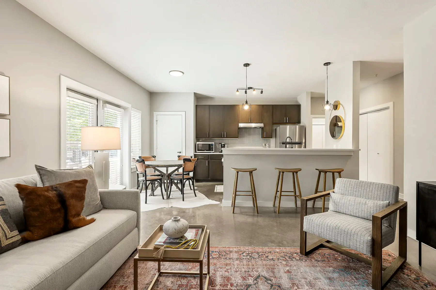 Sala de estar y cocina modernas de concepto abierto en tonos neutros, con un sofá, un sillón, un juego de comedor de madera y lámparas colgantes sobre una isla de cocina.
