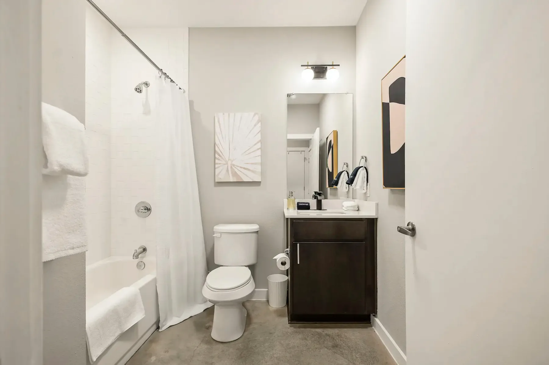 Baño moderno con bañera y ducha combinadas, cortina blanca, inodoro, mueble de lavabo oscuro y arte abstracto en la pared sobre el toallero. Decoración minimalista.