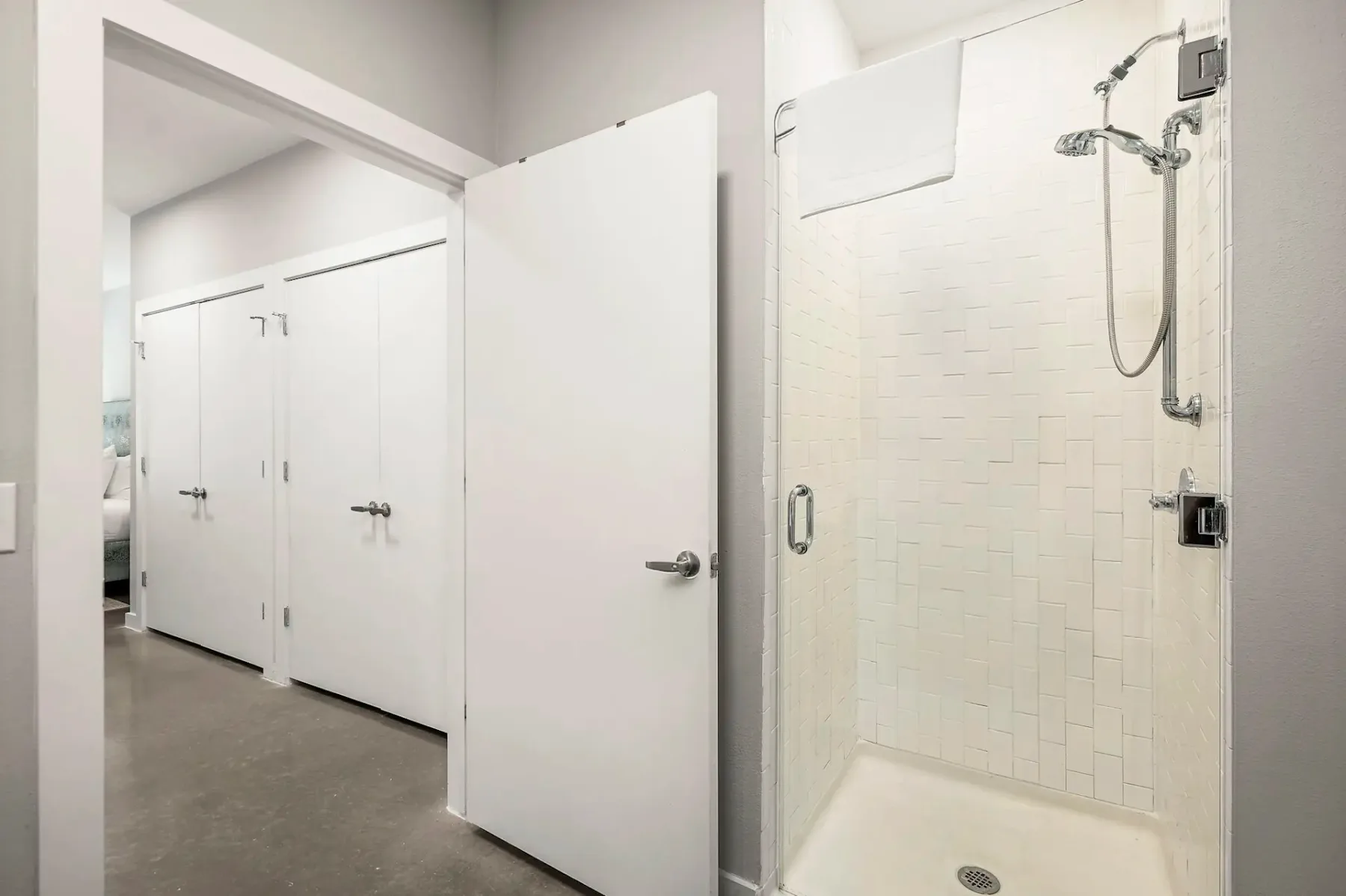 Baño minimalista con una ducha cerrada con mampara de vidrio y una toalla colgada encima. Las puertas abiertas dejan ver armarios y un dormitorio al fondo.