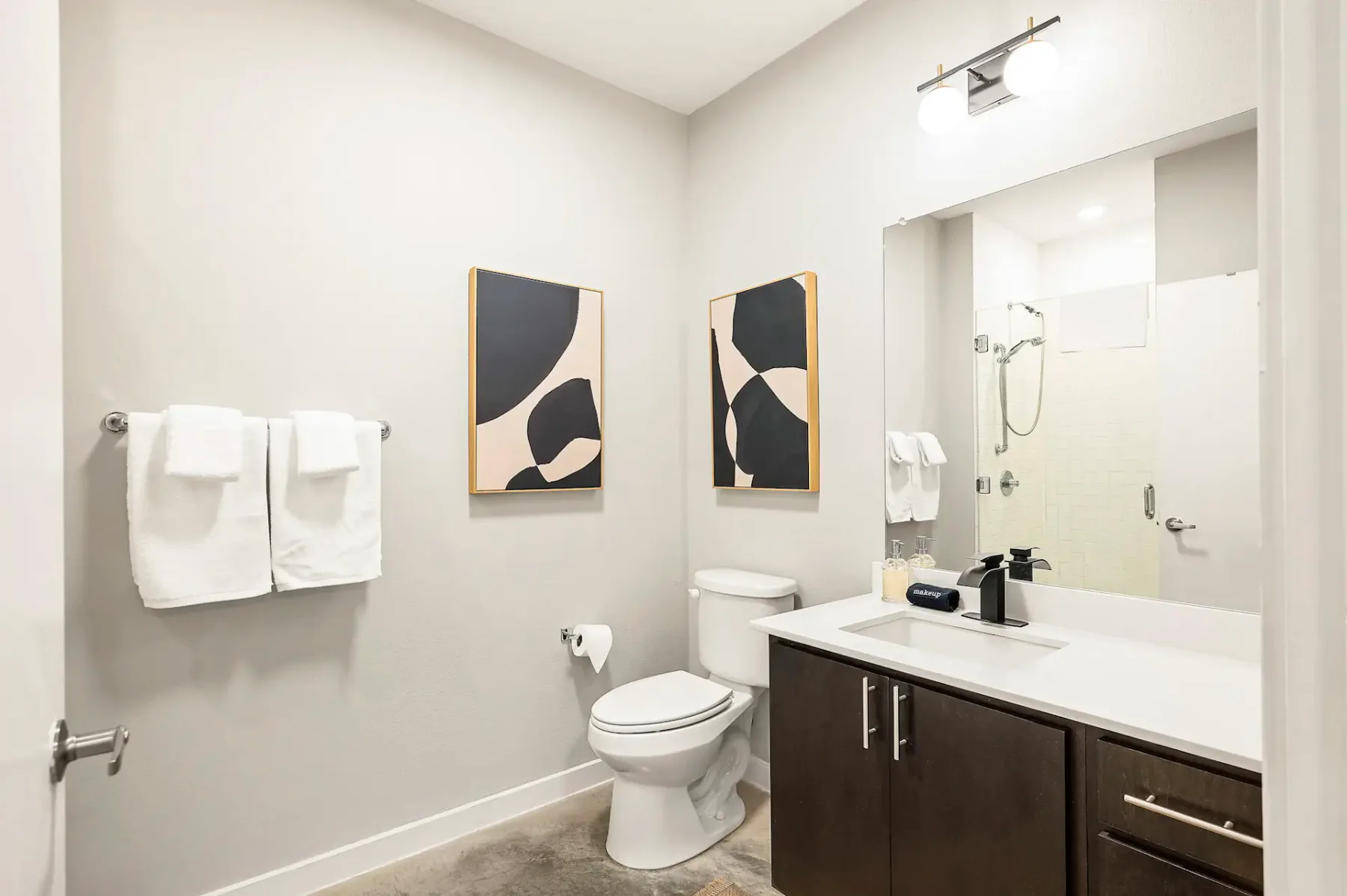 Baño moderno con arte abstracto, toallas blancas, un inodoro y un mueble de lavabo de madera oscura con un espejo y una lámpara encima.
