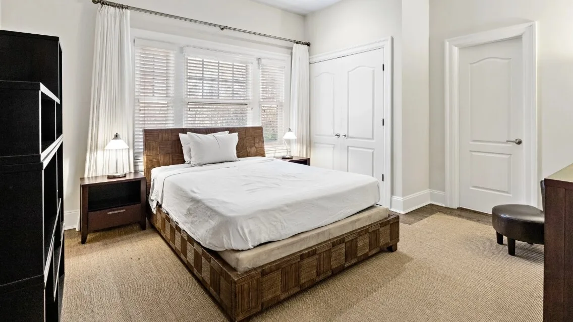 Dormitorio con una cama bajo una ventana grande y cortinas blancas.