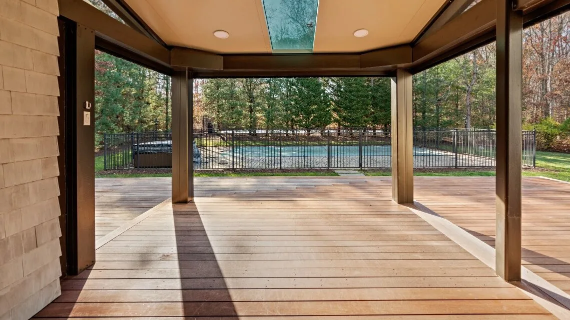 Una terraza de madera cubierta da a una zona de piscina cercada entre árboles.