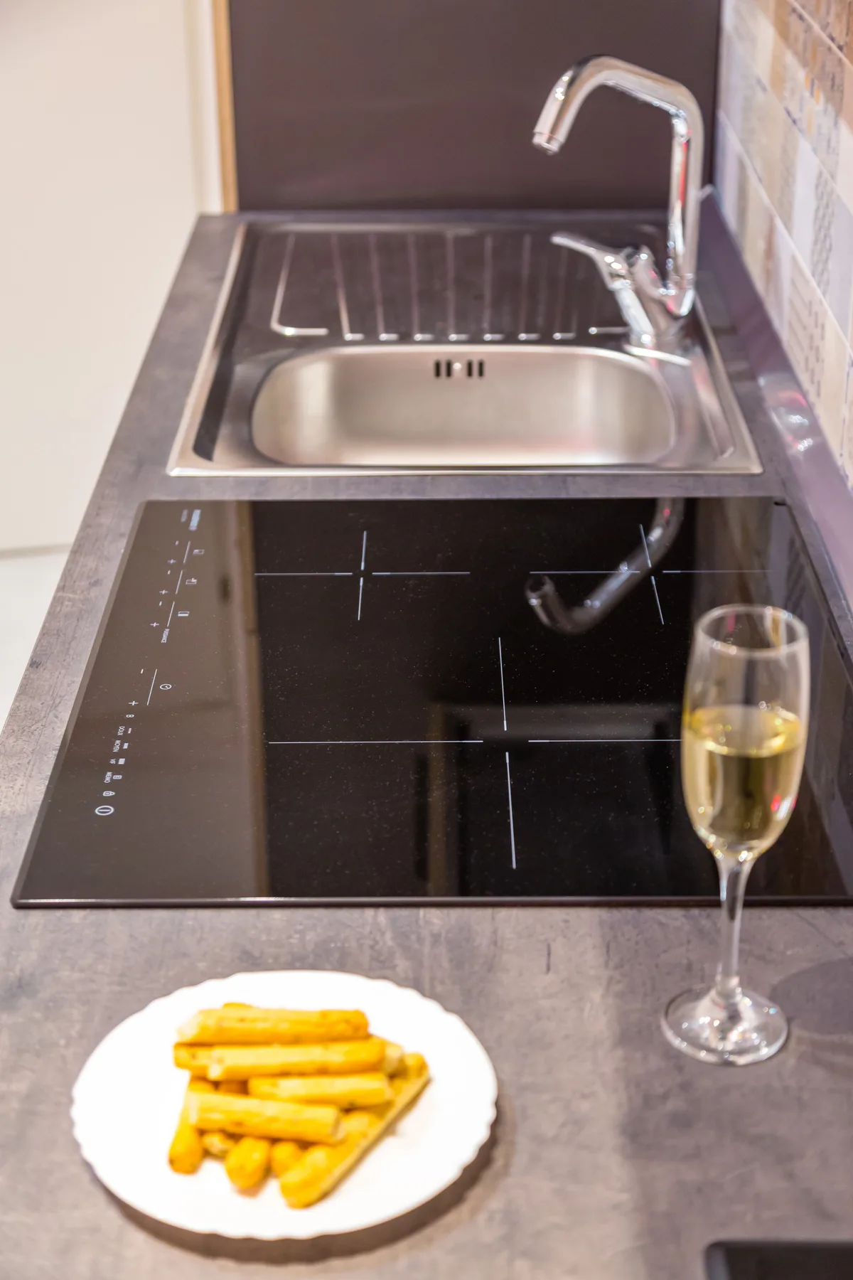 Una encimera de cocina compacta con fregadero, placa de cocción, papas fritas y una copa de vino.