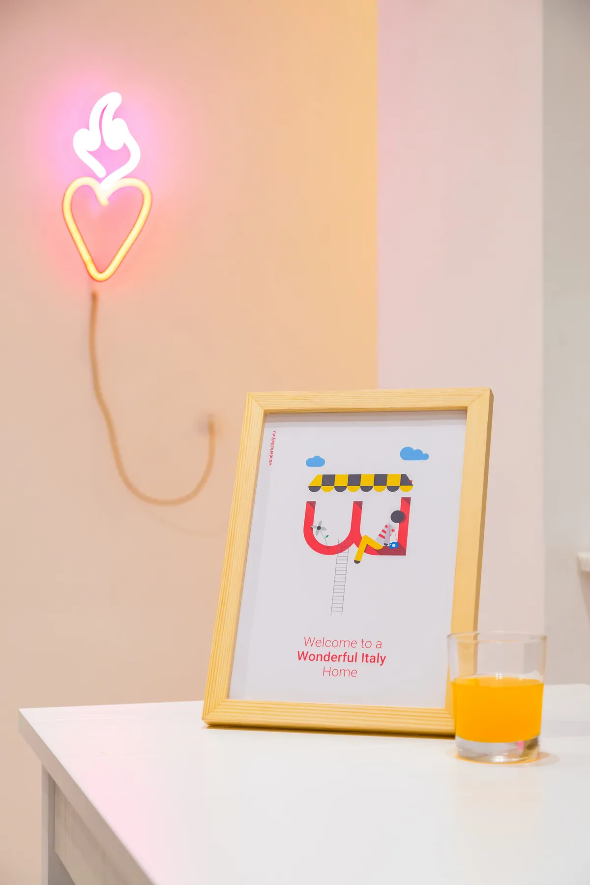 Un cartel de bienvenida enmarcado y un vaso de zumo de naranja están sobre una mesa blanca, bajo una luz de corazón.