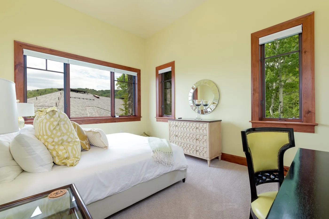 Un dormitorio luminoso con una cama, una cómoda y grandes ventanas con vistas a la vegetación.