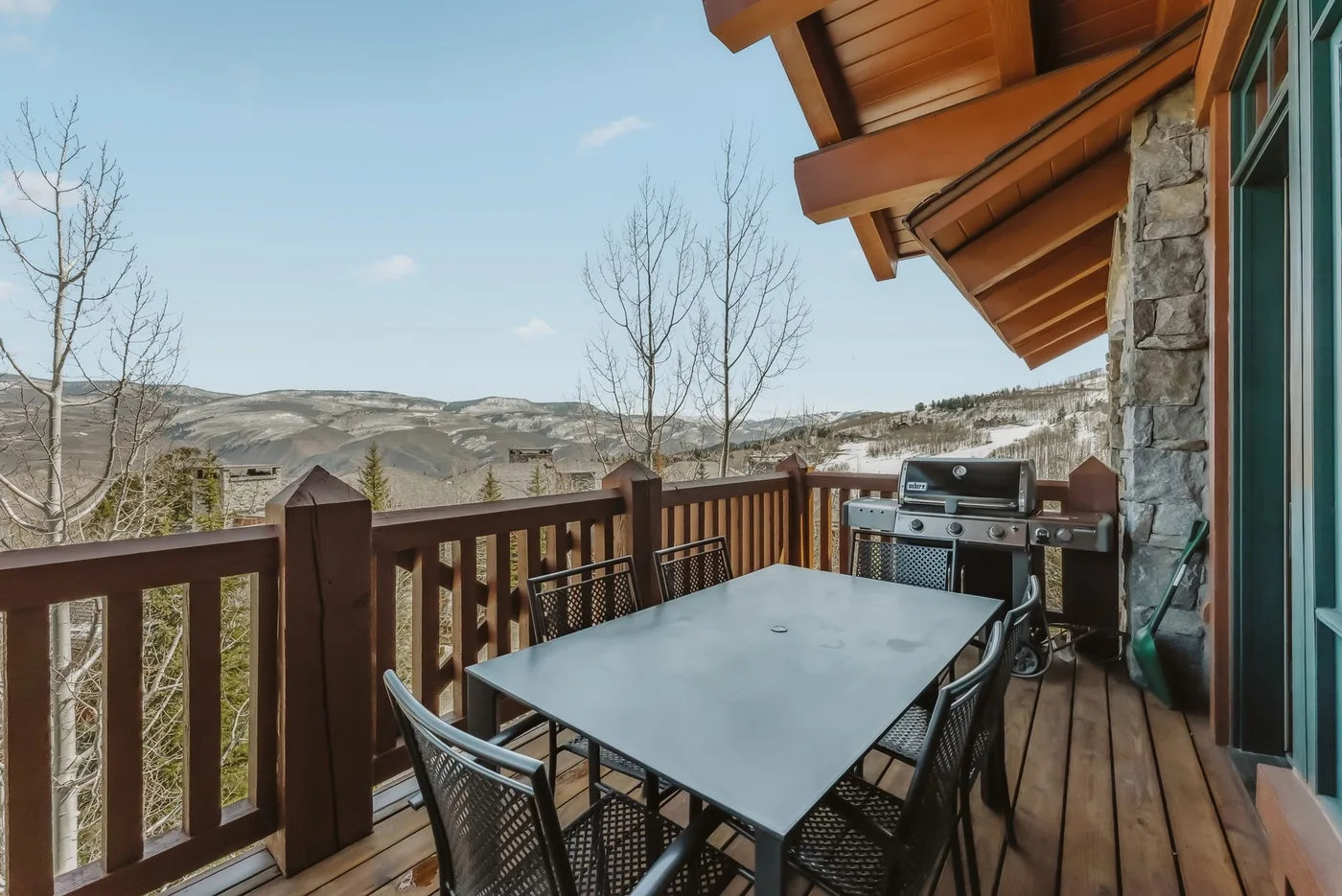 Una terraza de madera con mesa de comedor y parrilla da a un paisaje montañoso.