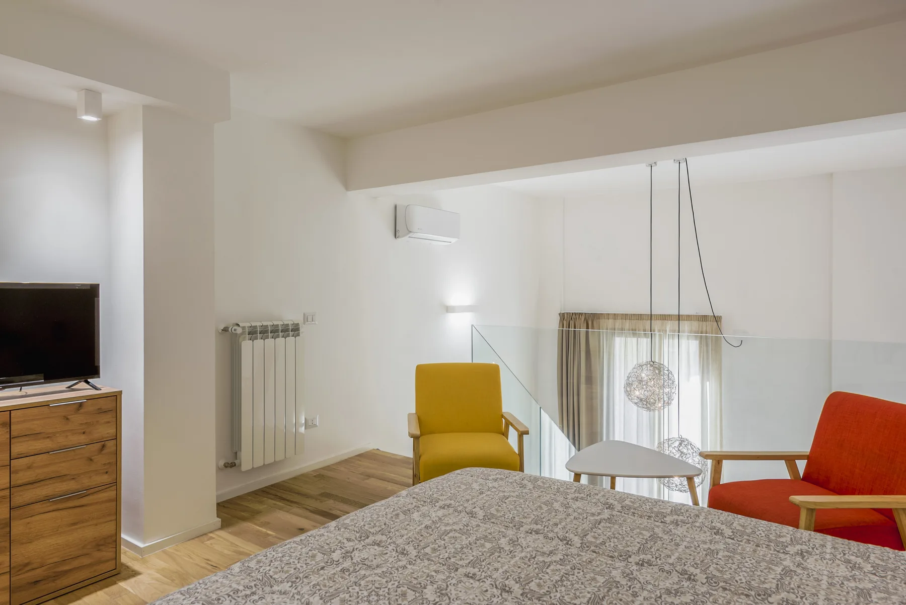A bright living room with a bed in the foreground and colorful seating by the window.