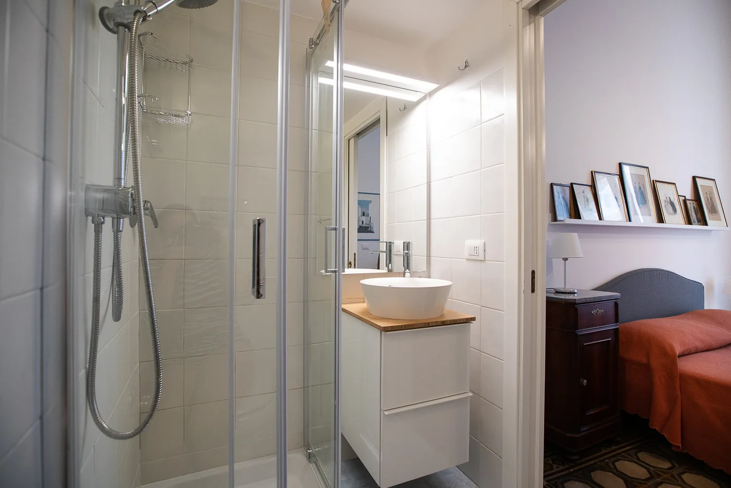 Un baño alicatado con ducha de cristal y lavabo blanco junto a un dormitorio.